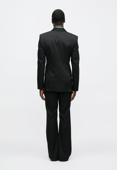 Čierny oblek s ostrými ramenami, zúženými klopami a nohavicami s širokými nohavicami. Výrazné detaily zahŕňajú ozdobné stehy na zadnej strane.