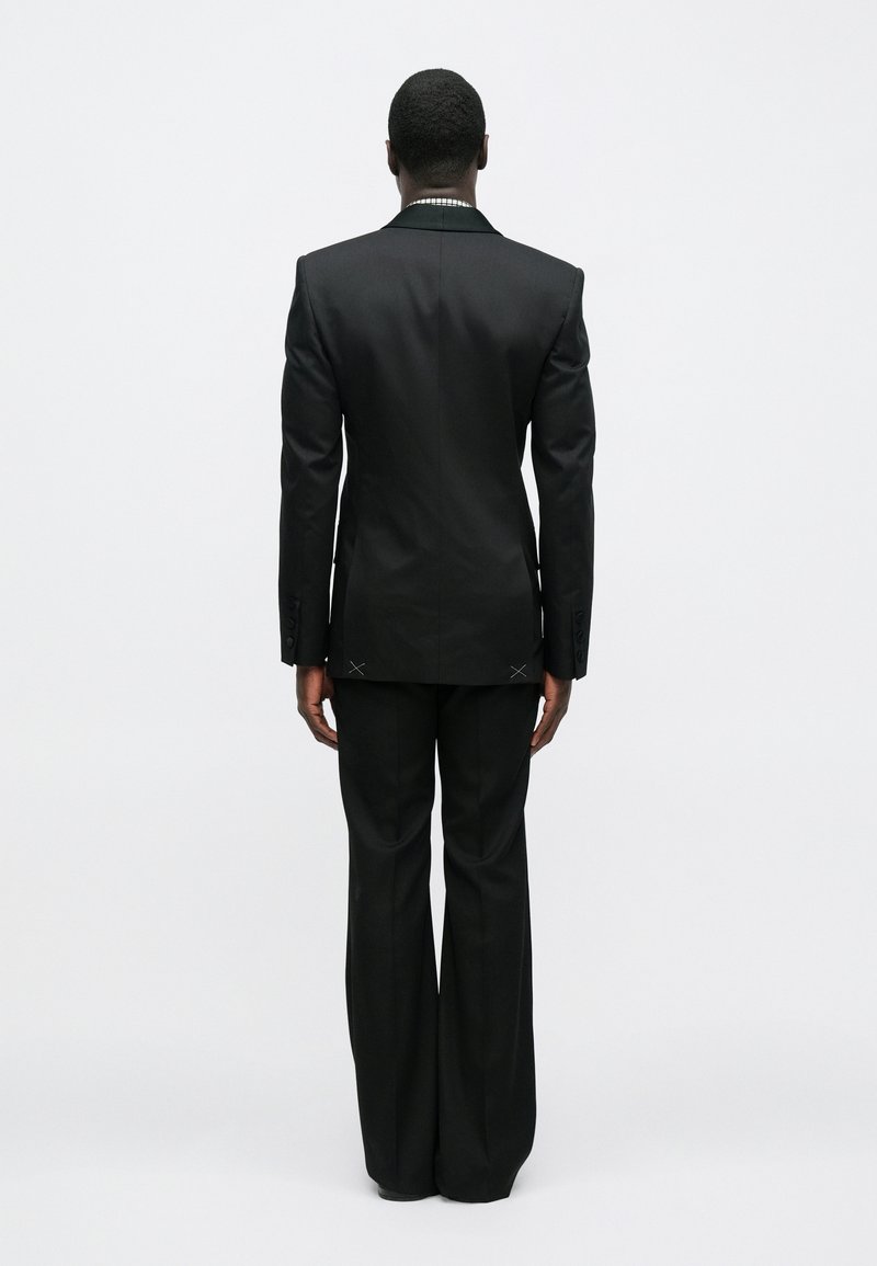 Costume noir avec épaules marquées, revers effilés et pantalons à jambes larges. Les détails notables incluent des accents de couture à l'arrière.