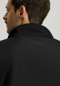 Czarny pulower z wysokim kołnierzem, wykonany z teksturowanej tkaniny, z małą naszywką logo na plecach oraz widoczną srebrną kolczykiem w uchu modelki.