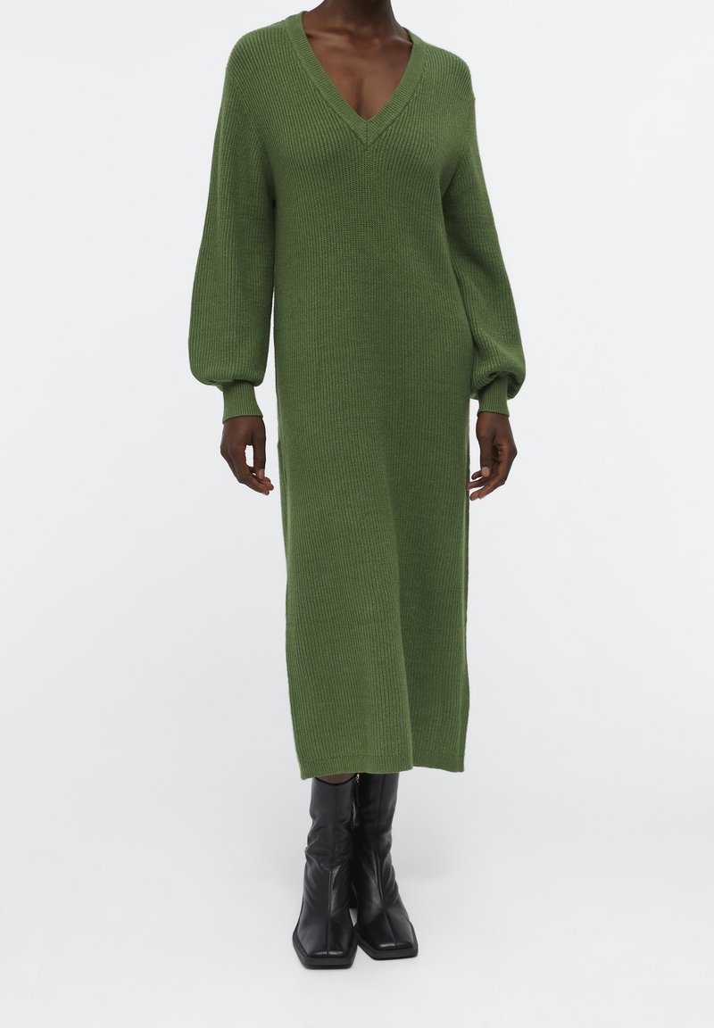 Personne à la peau foncée portant une longue robe tricotée vert olive avec des manches ballon et des bottes noires à bout carré sur fond blanc.