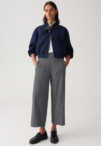 Giacca bomber blu navy con chiusura a zip, abbinata a pantaloni larghi grigi. Sciarpa trasparente a fantasia attorno al collo e scarpe nere.