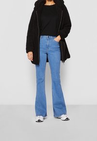 Abrigo con capucha forrado de felpa negra, camiseta negra, vaqueros acampanados azules, zapatillas blancas. Detalles de cremallera en el abrigo, ajuste informal en general.