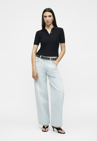 Γυναίκα που φοράει μαύρο κοντομάνικο πόλο μπλουζάκι, ανοιχτό μπλε τζιν με φαρδιά πόδια και μαύρη ζώνη, καθώς και μαύρα σανδάλια με τακούνι, στέκεται με το ένα χέρι στην τσέπη.
