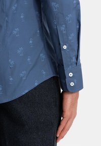 Camisa azul con un ligero patrón en relieve de flamencos y palmeras, con puños abotonados, fabricada en un tejido suave. Pantalones oscuros de textura.