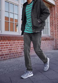 Giacca di jeans grigio scuro sopra una t-shirt a righe verdi e blu navy, abbinata a sneakers grigie. Camminando su un marciapiede texturizzato vicino a muri di mattoni.