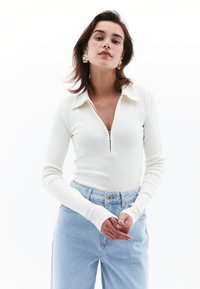 Pitkähihaisessa ribbipaidassa on off-white väri ja vetoketjukaulus. Se on yhdistetty korkeavyötäröisiin vaaleansinihin farkkuihin, jotka korostavat rentoa istuvuutta ja minimalistista ilmettä.