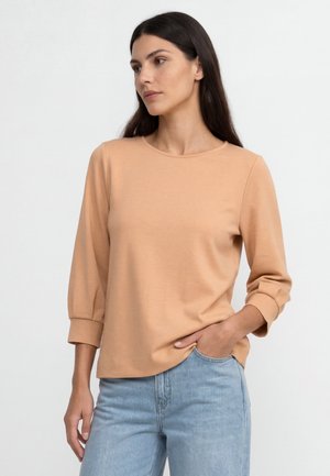 Femme aux longs cheveux foncés portant un haut beige à manches longues et un jean bleu clair, debout avec une main dans la poche sur fond blanc.