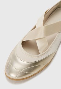 Scarpe piatte metalliche oro con punta arrotondata, caratterizzate da fasce incrociate in tessuto. Texture liscia con dettagli di cucitura lungo la parte superiore.