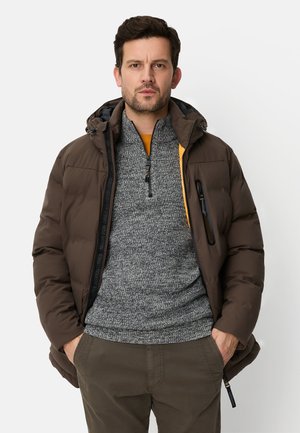 Braune Steppjacke mit Kapuze, vorne mit Reißverschluss und seitlichen Taschen. Trägt man über einem grauen Strickpullover und dunklen Hosen.