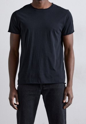 Mand iført en ensfarvet sort t-shirt med korte ærmer og sorte bukser, der står foran en ensfarvet lyse baggrund.