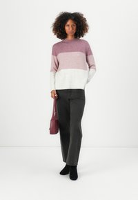 Farbblockierter Strickpullover in verschiedenen Lila- und Weißtönen, mit hohem Kragen und gerippten Bündchen, kombiniert mit hochtaillierten schwarzen Jeans.