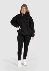 Zwarte oversized hoodie met voorzak en logodetail, gecombineerd met zwarte leggings en witte sportschoenen. Soepele stof, casual pasvorm.