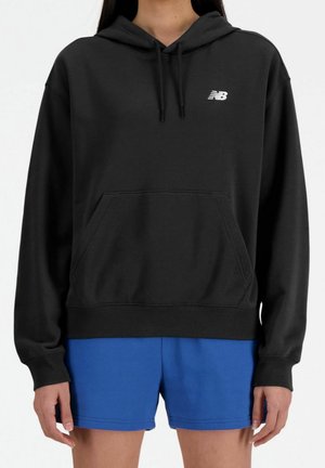 Person wearing a black New Balance hoodie with front pocket and blue shorts, standing against a plain background.