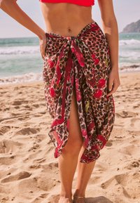 Paréo imprimé léopard avec des accents floraux rouges, en tissu léger, noué à la taille, doté d'une fente, porté sur une plage.