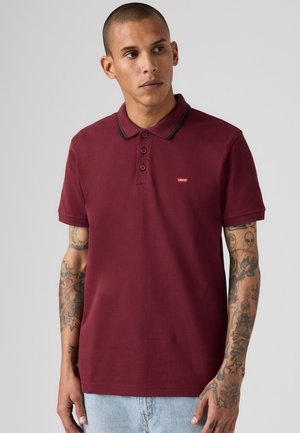 Homme aux cheveux courts et aux bras tatoués portant un polo Levi's bordeaux avec des rayures noires au col et un jean bleu clair.