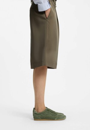 Person iført olivengrønne knælange shorts med hænderne i lommerne og matchende grønne ruskindssneakers, stående på en hvid baggrund.