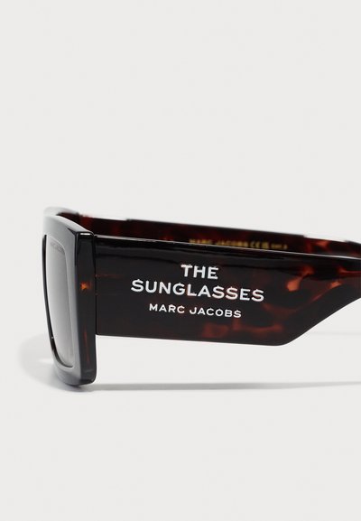 Ochelari de soare din tip tortoise, cu rame dreptunghiulare groase. Brațele prezintă text alb: "Ochelarii de soare" și "MARC JACOBS". Finisaj texturat.