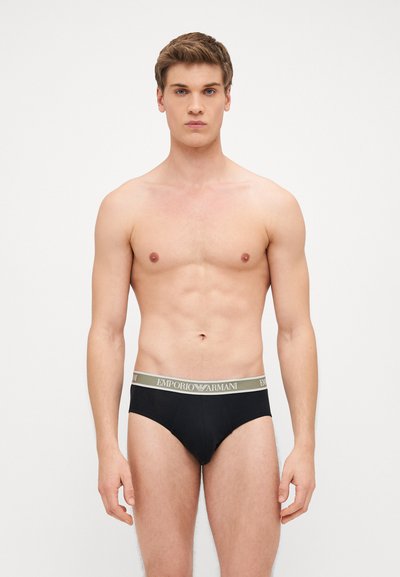 Slip neri da uomo realizzati in tessuto morbido con una vita caratterizzata da un logo verde che riporta la scritta "EMPORIO ARMANI." Il indossatore è in piedi con un'espressione neutra.