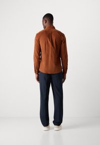 Cămașă lungă maro cu textură netedă, cu manșete cu nasturi și tiv curbat, purtată cu pantaloni negri închis și adidași albi.
