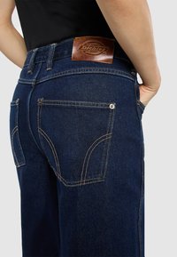 Persona che indossa jeans Dickies blu scuro con tasche posteriori visibili e un'etichetta in pelle del marchio, con una mano appoggiata nella tasca anteriore.