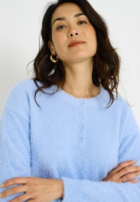 Jersey azul claro y peludo con cuello redondo, mangas largas y cuatro botones a juego en la parte delantera. Textura suave, ajuste informal.