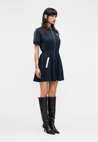 Φόρεμα σε ναυτικό μπλε με κοντά μανίκια, φερμουάρ στο μπροστινό μέρος, ελαστική μέση, λευκές ρίγες στο πλάι, με φουσκωτή φούστα, συνδυασμένο με μαύρες μπότες μέχρι το γόνατο.