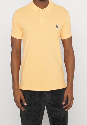 Mann trägt ein hellgelbes kurzärmeliges Polohemd mit kleinem Zebra-Logo auf der linken Brust und schwarze, strukturierte Hose.