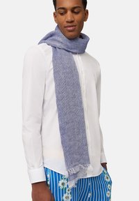 Sciarpa blu intrecciata con una finitura testurizzata, caratterizzata da bordi frangiati, indossata su una camicia bianca, abbinata a pantaloni a strisce con stampa floreale.