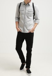 Chemise en jean gris à manches longues avec deux poches poitrine, associée à un pantalon noir et des chaussures noires sans lacets. Bretelles noires réglables.