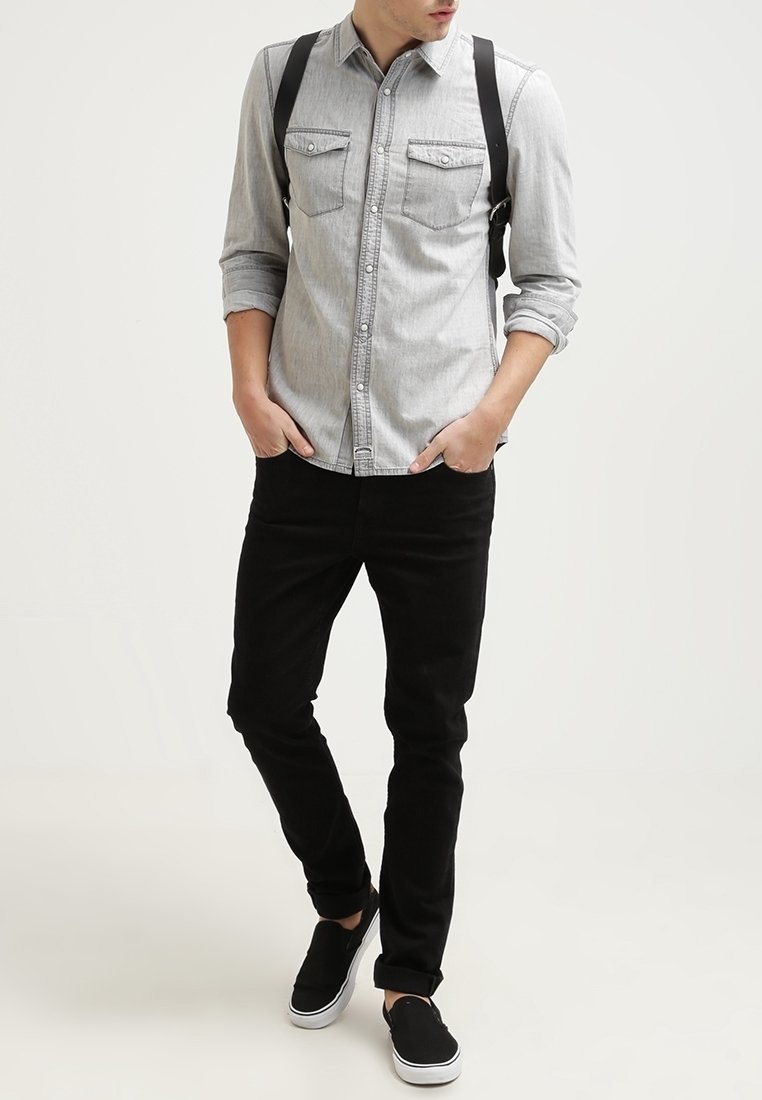 Chemise en jean gris à manches longues avec deux poches poitrine, associée à un pantalon noir et des chaussures noires sans lacets. Bretelles noires réglables.