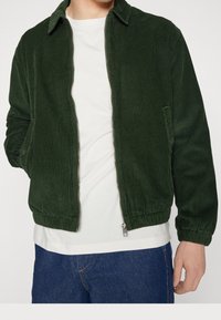 Donkergroene corduroyjas met een kraag, ritssluiting, zijzak en elastische zoom. Gedragen over een wit shirt en blauwe spijkerbroek.