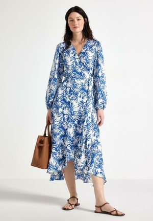 Donna con vestito bianco a maniche lunghe con stampa floreale blu, che tiene una borsa marrone, in piedi a piedi nudi su sfondo bianco.