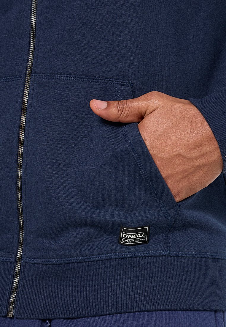 Granatowy rozpinany hoodie wykonany z miękkiego materiału, z przednią kieszenią, ściągaczami przy mankietach oraz małą metką marki na dolnym brzegu.