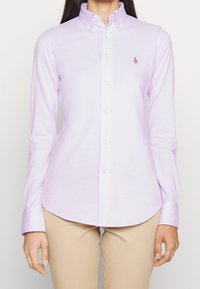 Camisa de manga larga de color lila claro con cuello abotonado, que presenta un pequeño logo bordado en el pecho y botones perlados.