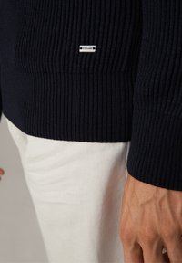 Maglione navy a coste con design aderente, tessuto strutturato e un piccolo accent logo in metallo. Indossato sopra pantaloni bianchi.