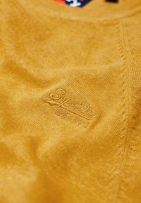 Maglione a maglia giallo senape con logo "SuperDry" in rilievo, texture dettagliata e rifiniture a coste su polsini e orlo.