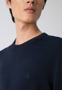 Marineblauer gestrickter Pullover mit Rundhalsausschnitt. Besitzt eine strukturierte Oberfläche und ein dezentes, auf der Brust gesticktes Logo.