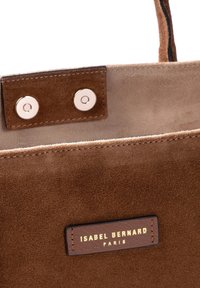 Sac fourre-tout en daim marron à texture lisse, doté d'une étiquette en cuir avec un texte doré embossé. Fermeture sécurisée par des boutons-pression argentés.