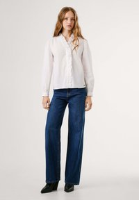 Giovane donna con lunghi capelli ondulati, che indossa una blusa bianca a volant, jeans a gamba larga blu e stivaletti neri con tacco, in piedi su uno sfondo semplice.