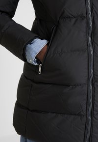 Doudoune noire avec des coutures horizontales, une fermeture éclair devant et des poches latérales. Une chemise en denim clair est visible sous la doudoune.