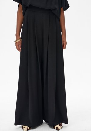 Kobieta nosi czarne spodnie z wysokim stanem o szerokich nogawkach z plisami, zestawione z butami w krowi wzór na zaostrzonych czubkach oraz złotymi bransoletkami na lewej ręce.