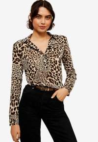 Blusa a maniche lunghe con print leopardato in tonalità beige e nero. Presenta un colletto e tre bottoni sul petto. Abbinata a pantaloni neri.