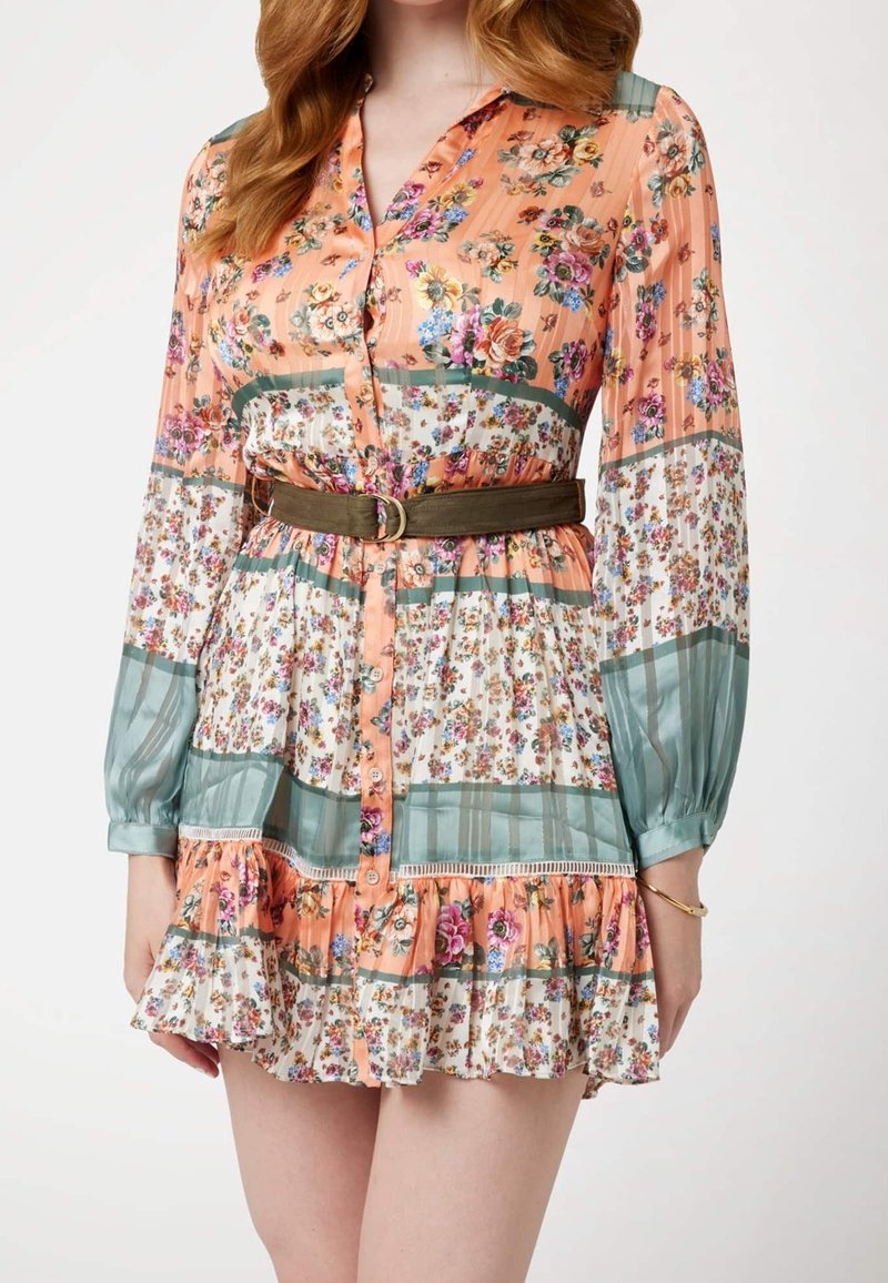 Femme portant une mini-robe à manches longues à motif floral avec une ceinture marron et un ourlet à volants.