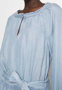 Ljusblå denimklänning med volangkrage, nyckelhålsutskärning och självknuten midja. Har långa ärmar och en avslappnad passform.