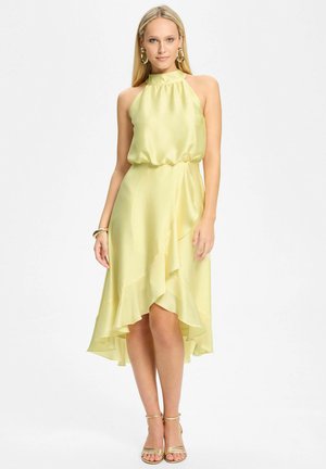 Kobieta w bezrękawną bladożółtą satynową sukienkę z wysokim dekoltem i asymetrycznym falbanowym dołem, w połączeniu ze złotymi sandałami i kolczykami kołami.