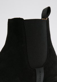 Bottine en daim noir avec panneau latéral élastique, finition texturée et languette de tirage. Présente une silhouette épurée et des détails de couture subtils.