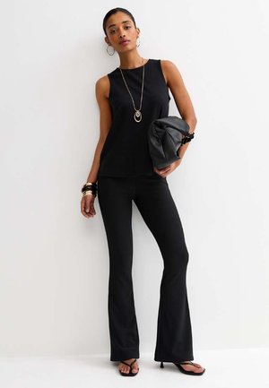 Kobieta ubrana w czarny bezrękawnik, czarne rozszerzane spodnie, sandały, złotą biżuterię, trzyma czarną kopertówkę.