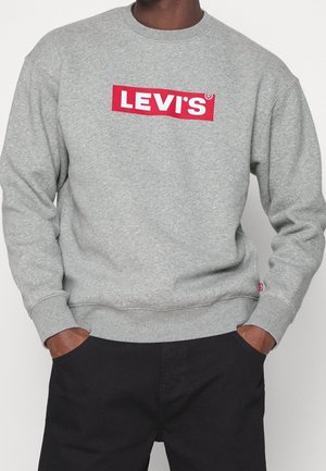 Person iført en grå Levi’s sweatshirt med rød og hvidt logo, hænder i lommerne, kombineret med sorte bukser, foran en ensfarvet baggrund.