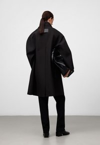 Cappotto oversize nero con colletto alto e una toppa posteriore rettangolare, abbinato a pantaloni neri e stivali alla caviglia su uno sfondo neutro.