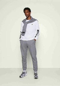 Jovem vestido com um moletom branco de mangas longas com texto preto nas mangas, calças de moletom cinzentas, um suéter cinzento caído sobre os ombros e ténis.